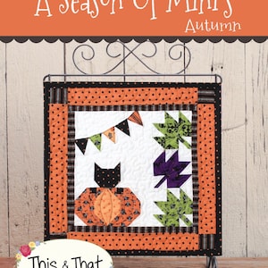 May include: A miniature quilt block with a black cat sitting on a pumpkin, surrounded by orange and black polka dot fabric, green and purple leaves, and a string of flags. The quilt block is framed with a black and white striped border and a black and orange polka dot border. The quilt block is hanging on a black metal stand. The quilt block is titled "A Season of Mini's Autumn" and is labeled "This & That Original Quilt Patterns by Sherri K. Falls #314".