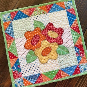 May include: A colorful patchwork quilt with a green border and a floral applique design. The applique features two flowers in shades of red, orange, and yellow with white polka dots.