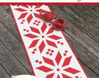 May include: A red and white table runner with a Scandinavian snowflake design. The runner is made of fabric with white polka dots on a red background. The runner is on a wooden surface with a red teapot and a red teacup.