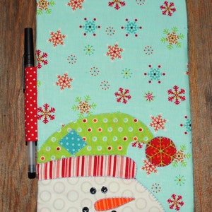 May include: A blue fabric notebook cover with a snowman design. The snowman has a green hat with white polka dots, a red and white striped hat, and a red pom-pom. The background is covered in snowflakes.