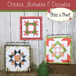 May include: Three quilt blocks, each with a different pattern, are displayed on a wooden surface. The blocks are made with red, green, white, and black fabrics. The blocks are titled "Bitty Barn Blocks" and "This & That".