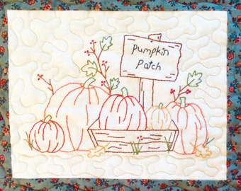Puede incluir: Tela blanca bordada con una escena de calabazas en una caja de madera. Las calabazas son naranja y amarillas. Hay un cartel que dice "Pumpkin Patch" en letras marrones.