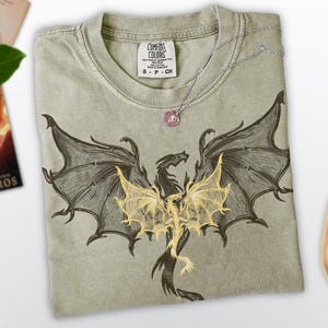 Op de afbeelding: Een lichtgroen T-shirt met een zwarte en gouden draakprint. De draak heeft zijn vleugels uitgespreid en kijkt naar voren. Het T-shirt heeft een label met de tekst "Comfort Colors".