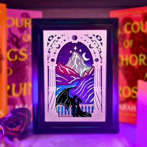May include: A rectangular black frame containing a multi-layered paper cut art piece. The scene depicts a silhouette of two figures, one with large wings, standing on a balcony before a mountain range under a night sky with stars and a crescent moon.