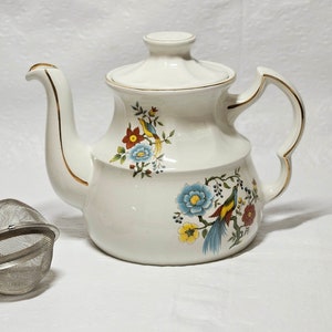 May include: White teapot with a gold trim and floral design featuring birds. The teapot has a lid and a handle. A silver tea strainer is in the foreground.