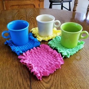 May include: Four colorful ceramic mugs with handles, each sitting on a handmade crocheted coaster in a different color: blue, yellow, green, and pink.