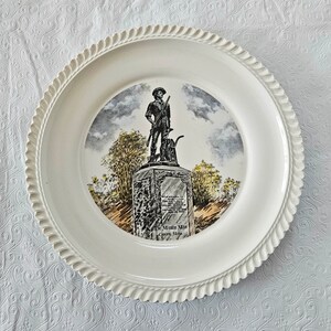 May include: White ceramic plate with a raised rim and a detailed image of a statue of a man with a rifle and a dog. The statue is on a pedestal with the text "The Minute Man Concord, Mass." below it.