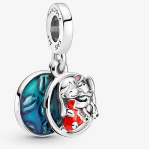 May include: A silver charm with a round pendant featuring a cartoon girl in a red dress hugging a cartoon animal. The charm has a blue and green marbled design on the back and the word "Pandora" is engraved on the top.