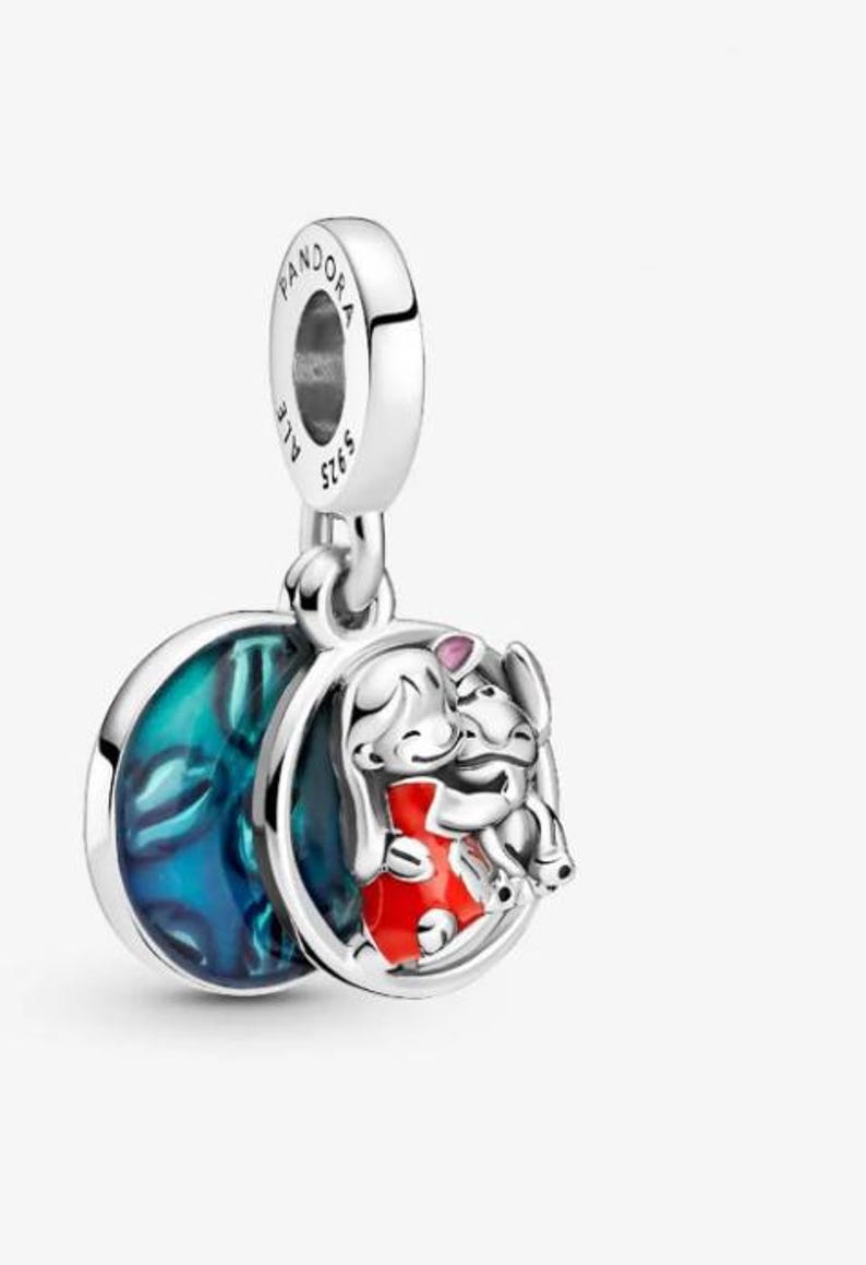 May include: A silver charm with a round pendant featuring a cartoon girl in a red dress hugging a cartoon animal. The charm has a blue and green marbled design on the back and the word "Pandora" is engraved on the top.