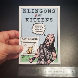 Op de afbeelding: Een stripboekachtige print getiteld "Klingons and Kittens" met een lichtblauwe achtergrond. De afbeelding toont een cartoon van een Klingon-krijger en een kattenreddingssc&egrave;ne. De tekst luidt "Misschien is vandaag een goede dag om te adopteren!"