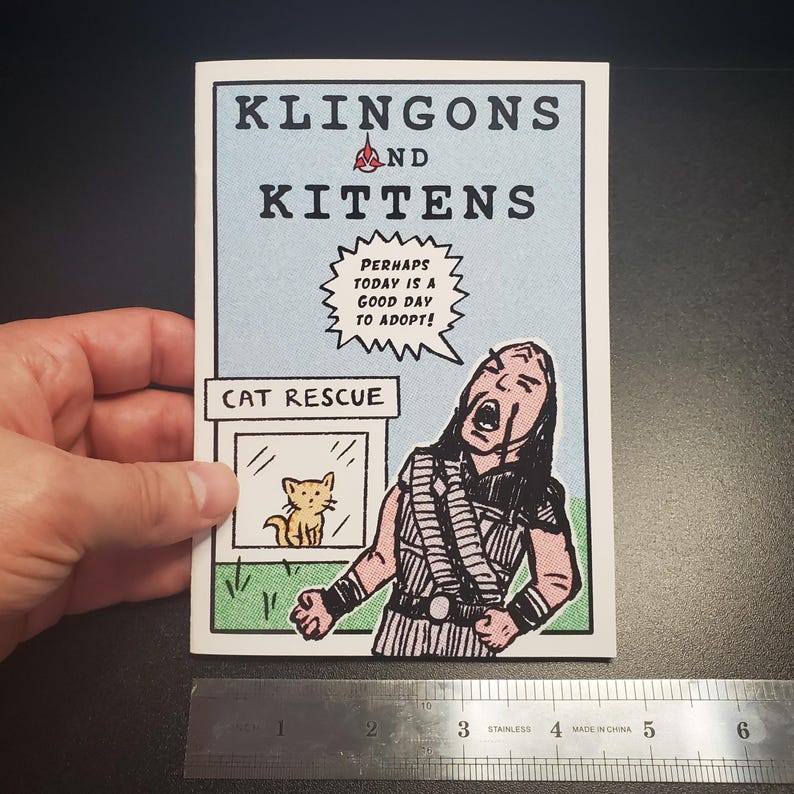 Op de afbeelding: Een stripboekachtige print getiteld "Klingons and Kittens" met een lichtblauwe achtergrond. De afbeelding toont een cartoon van een Klingon-krijger en een kattenreddingssc&egrave;ne. De tekst luidt "Misschien is vandaag een goede dag om te adopteren!"