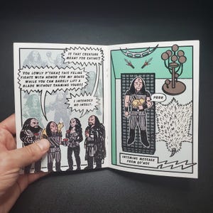 Op de afbeelding: Een stripboekspread met zwart-wit illustraties en tekstballonnen. De linkerpagina toont een groep personages met tekst, terwijl de rechterpagina een personage afbeeldt dat een kat vasthoudt. Tekst bevat "Incoming message from Qo'nos."