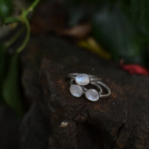 May include: Three silver rings with oval moonstone settings. The rings are stacked on top of each other on a dark brown textured surface.