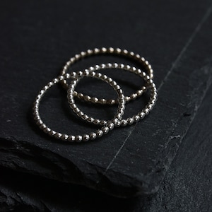 May include: Three silver beaded bracelets stacked on top of each other on a black slate surface.
