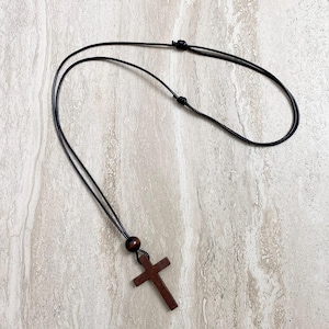 May include: A dark brown wooden cross pendant necklace with a black cord. The cross is a simple, classic design. The necklace is displayed on a light-colored, textured surface.
