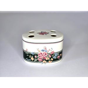 May include: A white ceramic toothbrush holder with a floral design. The holder has a dark green band with pink, blue, and yellow flowers. The top has four holes for toothbrushes and a small floral pattern.