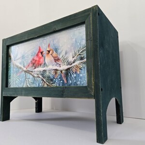 May include: A rectangular wooden box with a dark green distressed finish, featuring a central panel print of a male and female cardinal perched on a snow-covered pine branch. The box is elevated on four legs with arched cutouts between them.