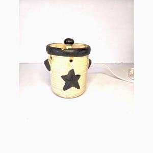 May include: Cream-colored ceramic electric wax warmer with a black star and rim. The warmer has a lid with a black knob, two small handles, and a power cord. The design is rustic and country-themed.