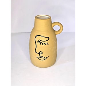 May include: Mustard-colored ceramic vase with a minimalist face design. The vase has a rounded body, a small handle, and a white interior. The face is drawn with black lines, featuring a closed eye, nose, and a smiling mouth.