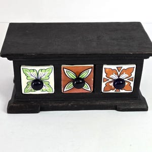 May include: A small, dark wooden box with three decorative drawers. Each drawer features a square, ceramic tile with a floral design in green, orange, and white, and a dark blue knob. The box has a rectangular shape and a slightly sloped top.