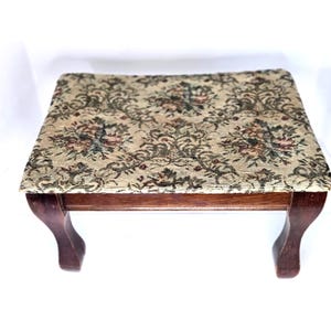 May include: Small, antique footstool with a rectangular top and curved legs. The top is upholstered with a floral tapestry in shades of beige, green, and burgundy. The wooden frame and legs are a dark brown color.