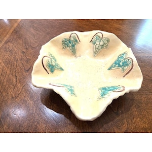 May include: A cream-colored, scalloped-edge ceramic dish with a hand-painted design of teal-colored leaves and brown stems. The dish is likely for decorative or serving purposes, showcasing a nature-inspired theme.