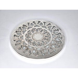 May include: A silver-colored, circular coaster with an intricate, openwork design. The coaster features a central floral pattern surrounded by a detailed, repeating motif. The edges are slightly raised, and the overall aesthetic is ornate and decorative.