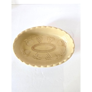 May include: An oval-shaped, cream-colored ceramic serving dish. The dish features embossed fruit and leaf designs, including grapes, strawberries, and a central oval. The rim has a scalloped edge.
