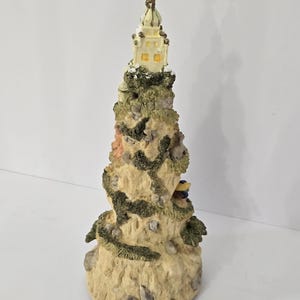 May include: A conical sculpture depicting a rocky mountain landscape with textured beige stone surfaces, patches of green moss, and a small building at the peak. A gold-colored star sits atop the structure, and a small figure is visible on a ledge.