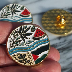 Può includere: Due spille in smalto dorate con sfondo bianco e disegni colorati. Una spilla presenta una pianta verde, onde blu e un'anguria rossa. L'altra spilla presenta un design simile con una bandiera palestinese.