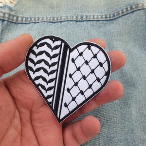 May include: A heart-shaped patch with a white background and black geometric patterns. One half features a chevron design, while the other has a grid pattern. The patch is held against a denim jacket.