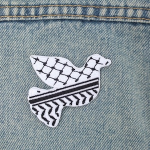 Pode incluir: Um patch de pomba branco e preto com um padrão geométrico, costurado em uma jaqueta jeans. O patch apresenta um design detalhado com um padrão xadrez nas asas e um padrão listrado no corpo.