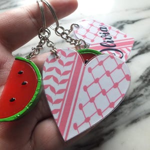 May include: Keyrings with a watermelon slice and heart-shaped charms. The watermelon slice is red with black seeds and a green border. The heart charms have pink and white patterns, with one bearing the name "Olivia".