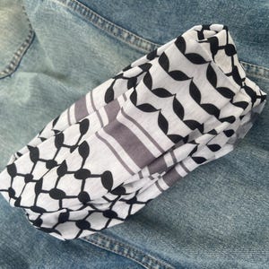 Pode incluir: Um lenço branco e preto com um padrão de folhas repetidas e riscas cinzentas, dobrado sobre uma jaqueta jeans azul. O padrão do lenço é uma mistura de formas geométricas e folhas estilizadas.