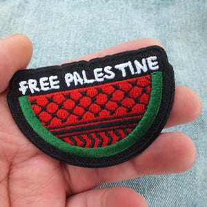 May include: Embroidered patch shaped like a watermelon slice, with the white text "FREE PALESTINE". The design incorporates red, black, and green colours, with a black border. The patch is held in a hand.