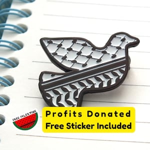 Könnte beinhalten: Eine taubenförmige Anstecknadel mit einem schwarz-weißen Muster, das einem traditionellen palästinensischen Design ähnelt, liegt auf einem linierten Notizbuch. Die Anstecknadel ist dunkelbraun mit weißen Details. Ein gelber Aufkleber mit Text ist ebenfalls sichtbar.