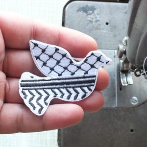 May include: A white dove patch with black geometric patterns, held in a hand near a sewing machine. The dove's wings and body feature distinct designs, including a crosshatch and a zigzag pattern. The patch is likely for clothing or fabric decoration.