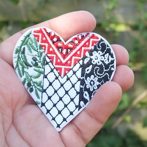 Può includere: Toppa ricamata a forma di cuore con un design patchwork. La toppa presenta sezioni di rami di ulivo, motivi geometrici e disegni floreali in bianco, rosso e nero. La toppa è tenuta in una mano.