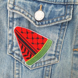 May include: A denim jacket with a watermelon patch on the pocket. The patch is a red watermelon slice with black seeds and a green rind. The jacket has a button closure and a silver button on the pocket.