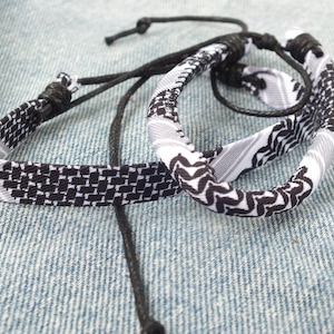 May include: Two black and white patterned bracelets with adjustable black cords. One bracelet is flat with a geometric design, and the other is a rounded bangle with a wavy pattern. The bracelets are displayed on a denim surface.