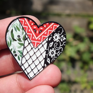 Puede incluir: Un pin de esmalte en forma de corazón con borde negro. El pin está dividido en cuatro secciones, cada una con un diseño diferente: ramas de olivo, un patrón geométrico rojo y blanco, un patrón floral en blanco y negro y una cuadrícula blanca.