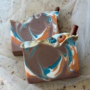 May include: Two rectangular bars of soap featuring a marbled swirl pattern in shades of brown, orange, white, and teal. The top of the soap is decorated with small, molded pumpkin shapes in blue and orange, along with a single brown cinnamon stick.