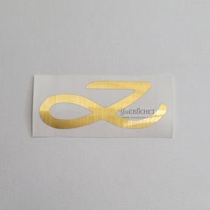 Puede incluir: Pegatina de vinilo dorada con la letra Z en una fuente cursiva. La pegatina tiene un acabado de metal cepillado y el texto "Yve Crochet" y "www.yvecrochet.com" está impreso debajo de la letra.