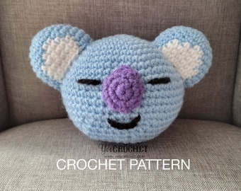 May include: A light blue crocheted stuffed animal shaped like a bear with white ears and a purple nose. The bear has a black mouth and closed eyes. The bear is sitting on a gray fabric surface. The text "Yecrochet" and "CROCHET PATTERN" is visible below the bear.