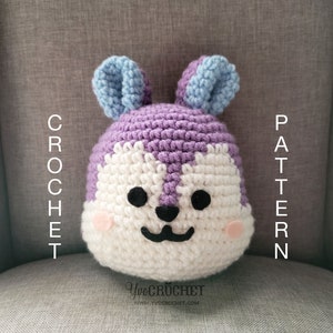 Könnte beinhalten: Gehäkeltes Plüschtier in Lila und Weiß mit blauen Ohren. Das Spielzeug hat schwarze Augen und eine schwarze Nase. Das Spielzeug sitzt auf einer grauen Stoffoberfläche. Der Text "CROCHET PATTERN" steht links und rechts im Bild.