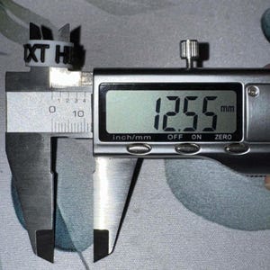 May include: A white ring with the letters "XT HI" is measured with a digital caliper. The caliper displays a measurement of 12.55 mm. The ring has several small, pointed extensions.