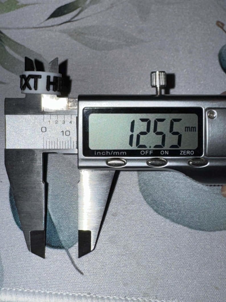 May include: A white ring with the letters "XT HI" is measured with a digital caliper. The caliper displays a measurement of 12.55 mm. The ring has several small, pointed extensions.