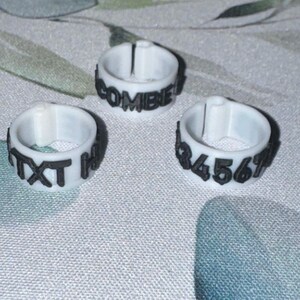 May include: Three white rings with black text. One ring reads "COMBIE!", another reads "TXT H", and the third reads "34567". The rings are made of a light-colored material and are set against a patterned background.