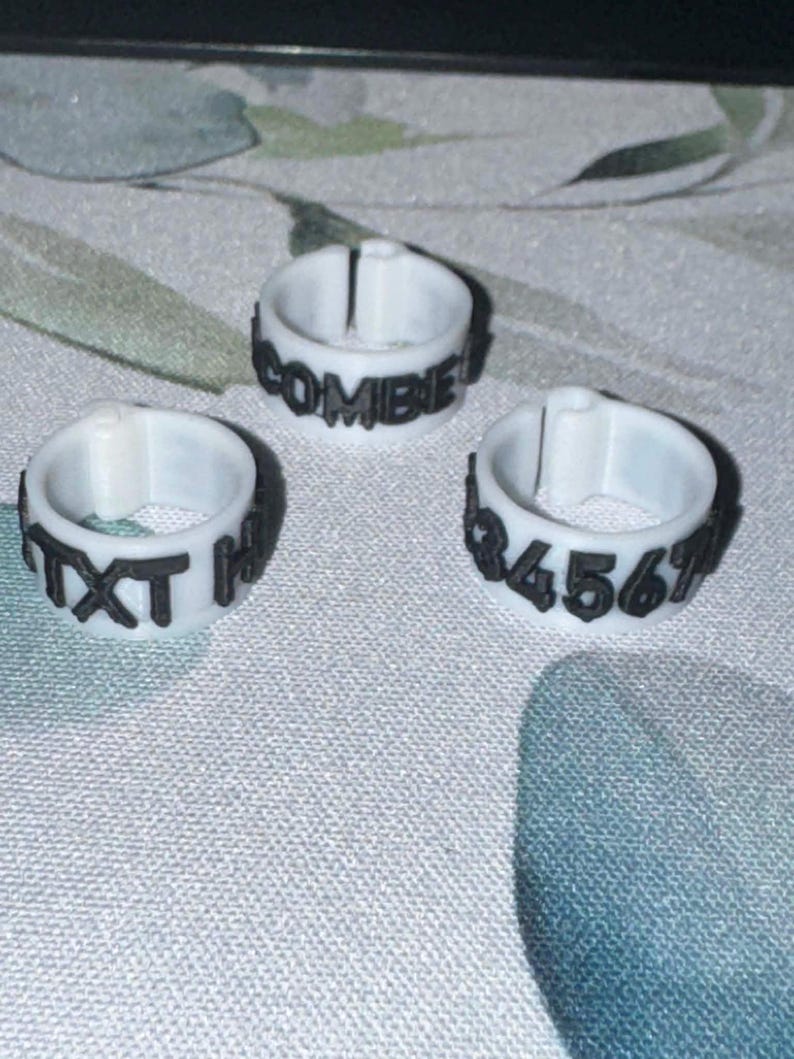May include: Three white rings with black text. One ring reads "COMBIE!", another reads "TXT H", and the third reads "34567". The rings are made of a light-colored material and are set against a patterned background.