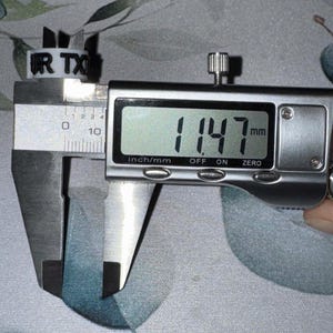 May include: A white plastic ring with the text "UR TX" is measured with a digital caliper. The caliper displays a measurement of 11.47 mm. The ring is positioned on a floral patterned surface.